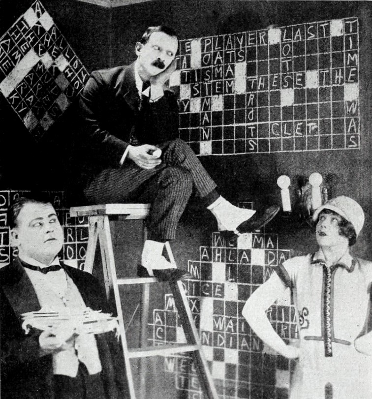 En komisk filmscene fra 1925 med tre personer foran store krydsord på væggene.